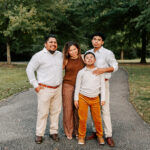 A mom and dad stand smiling with their two sons in white and brown outfits in a park trail at sunset