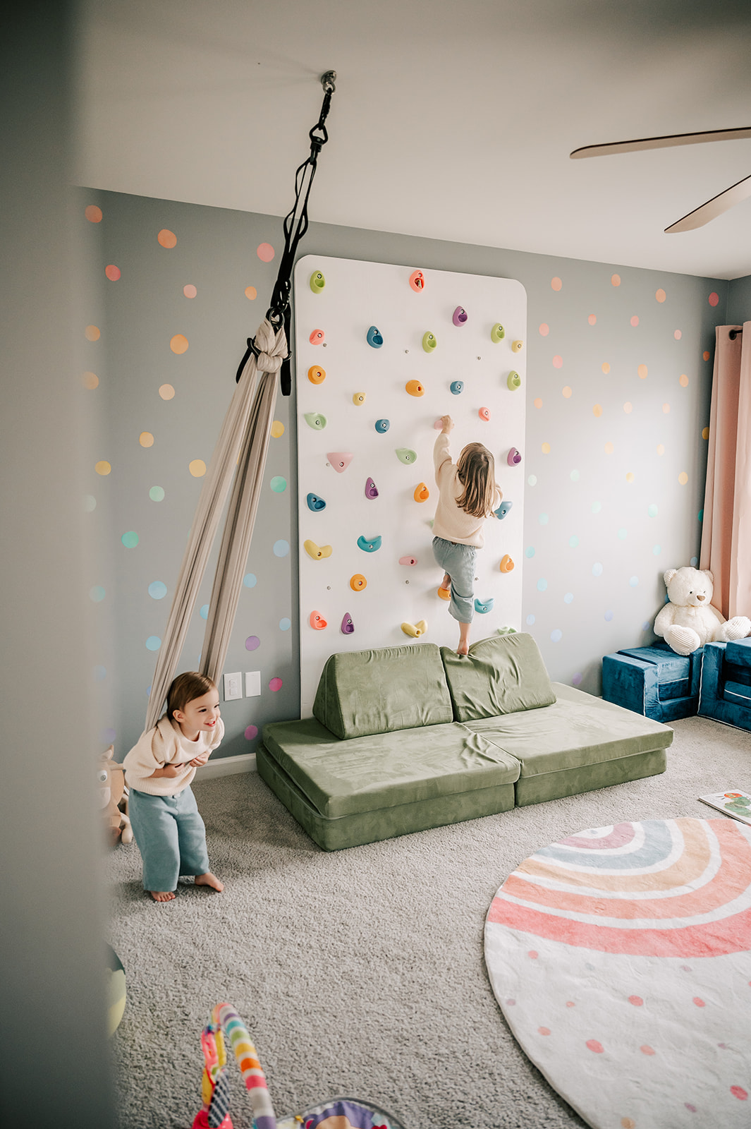 Toddler sisters play on a rock wall and silk swing in a play room