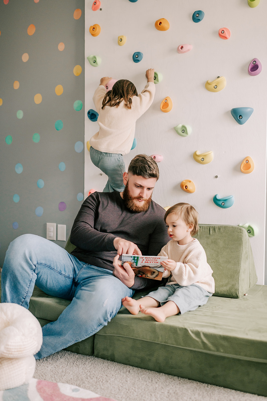 A dad in jeans and dark long sleeve reads a book with toddler daughter on a small couch as older daughter climbs rock twall behind them after visiting indoor playgrounds in Winston-Salem