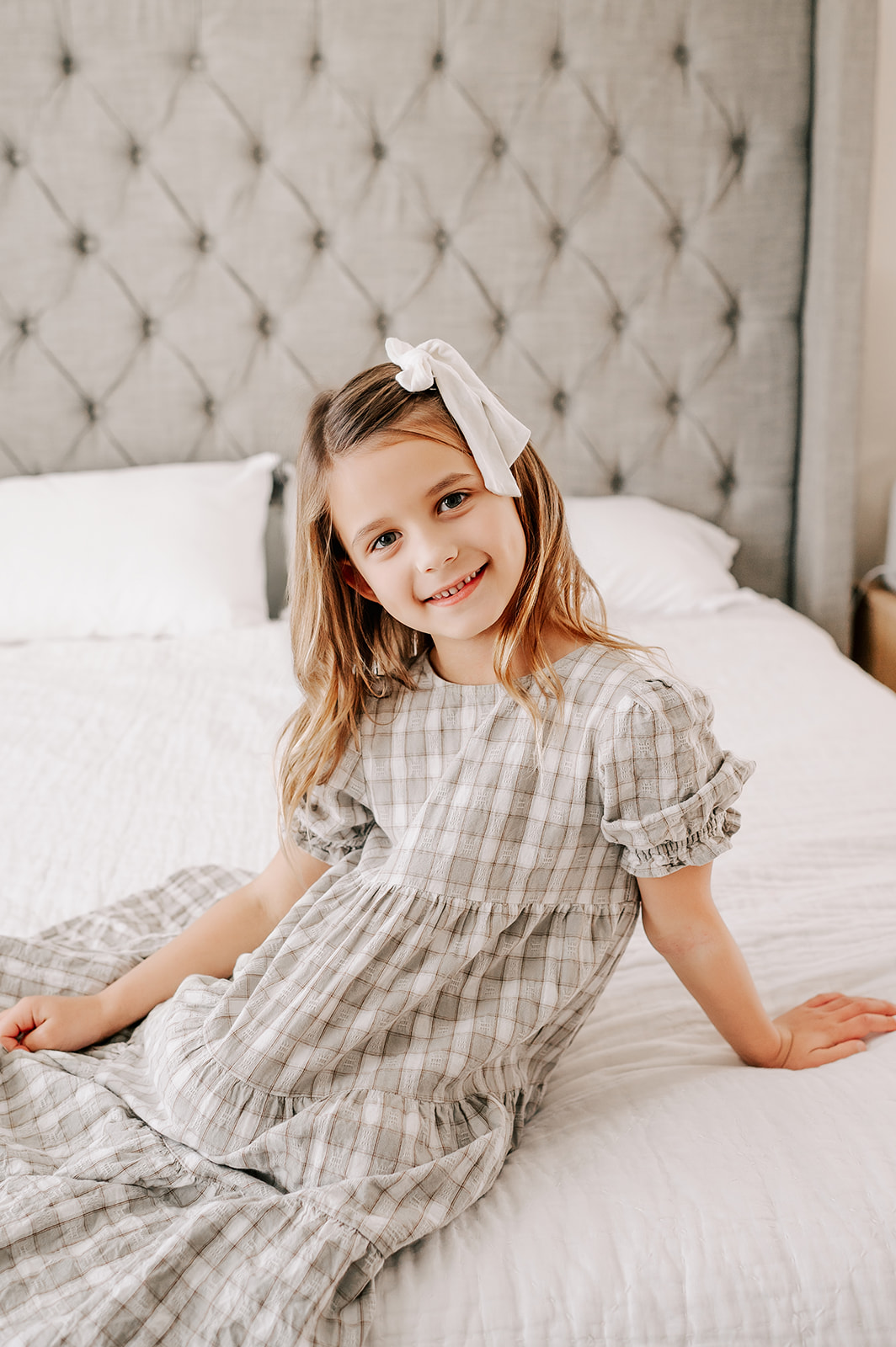 A young girl in a long sage dress and bow sits across a bed with a smile after some pediatric occupational therapy in Winston-Salem, NC
