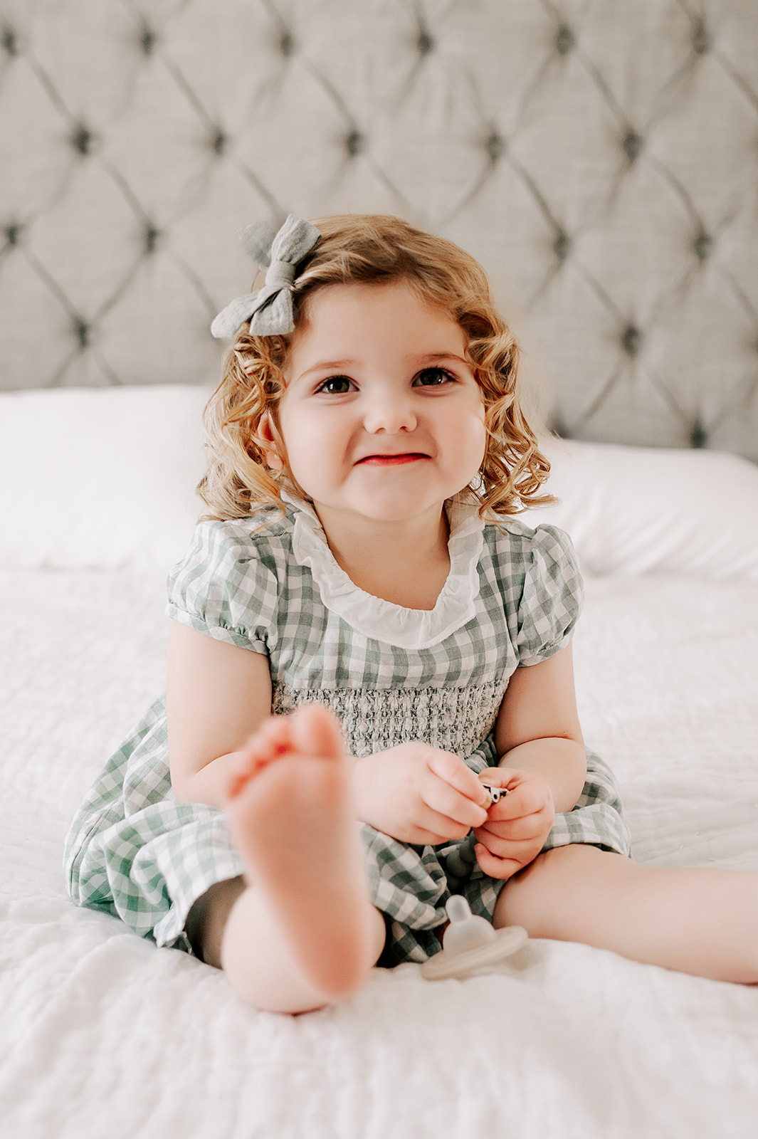 A happy toddler girl in a green dress sits on a bed with legs out after some pediatric occupational therapy in Winston-Salem, NC