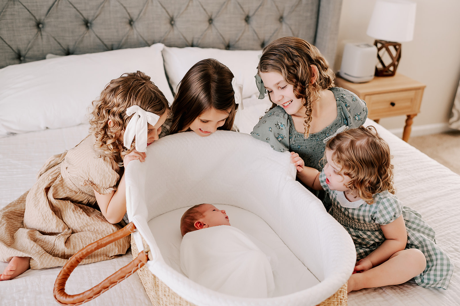 Four young sisters smile while sitting around their sleeping newborn sibling in a basket on a bed thanks to great pediatric occupational therapy in Winston-Salem, NC