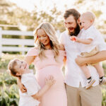 A mom and dad laugh with their happy young son in a garden at sunset with toddler in dad's arm before visiting Toys and Co.