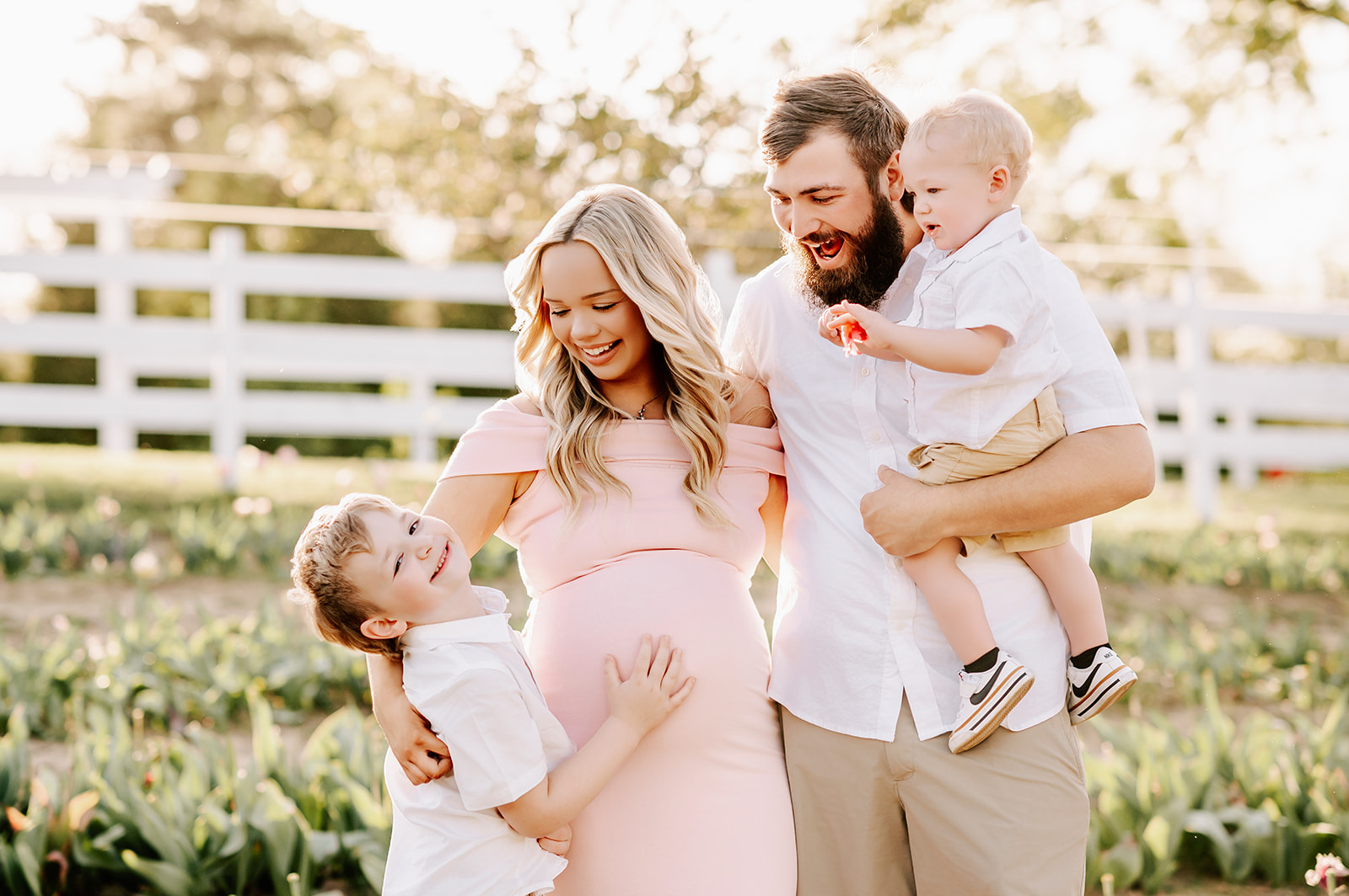 A mom and dad laugh with their happy young son in a garden at sunset with toddler in dad's arm before visiting Toys and Co.