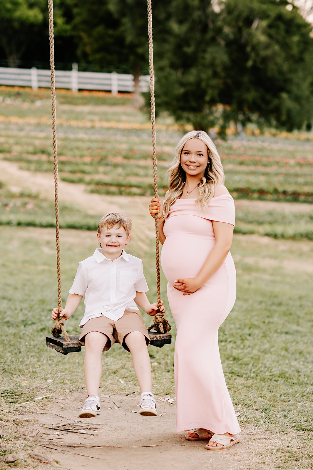 A happy pregnant mom in a pink maternity gown stands at a swing with her young son in a garden before visiting Toys and Co.