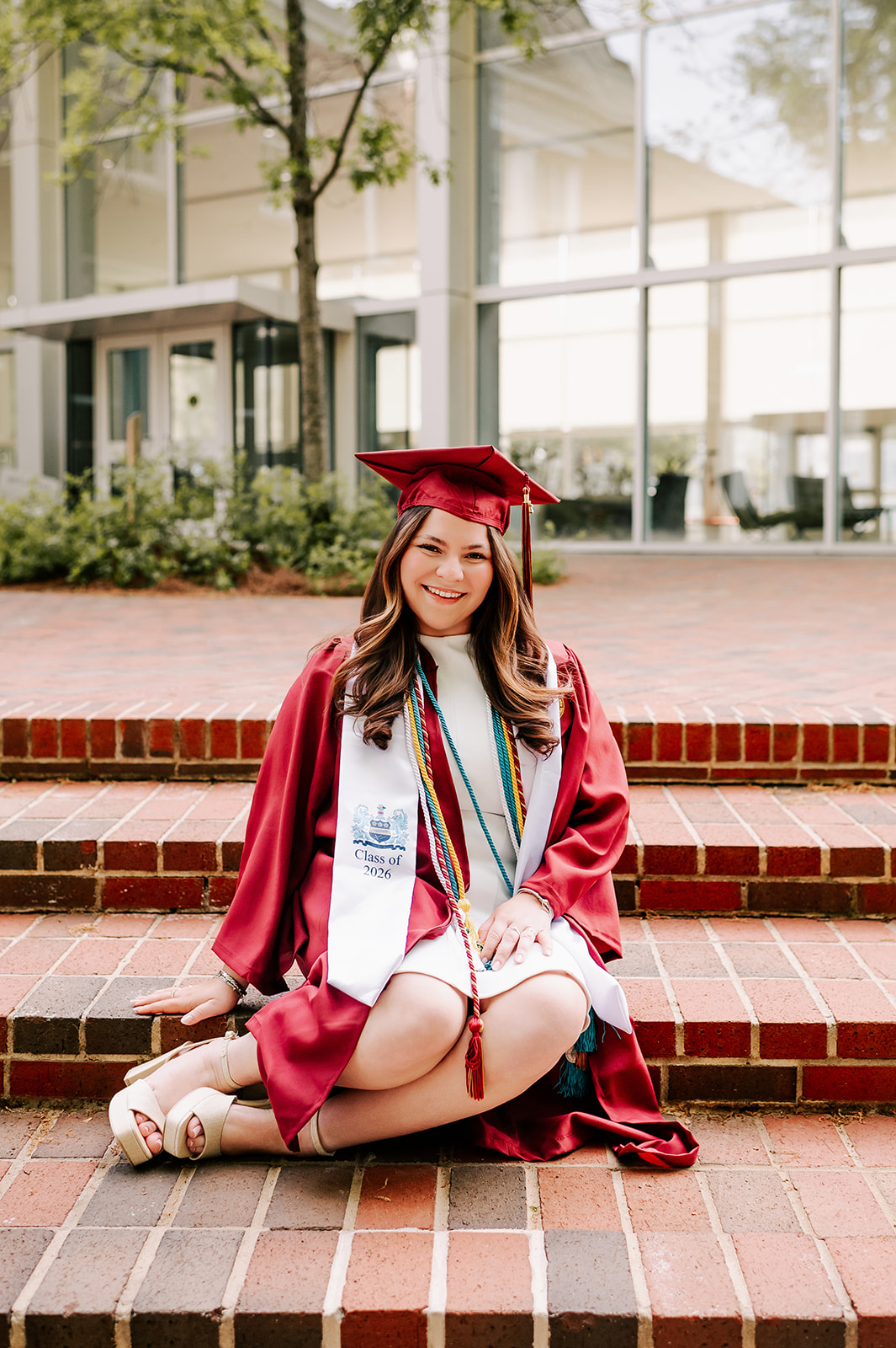 A smiling graduate sits on the steps of her building in red cap and gown