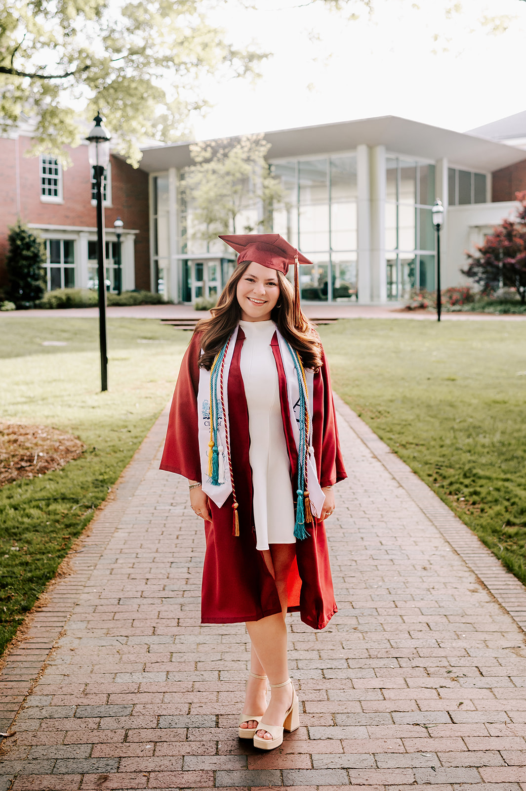 A happy graduate walks in the quad in her cap, gown and chords with a big smile on brick path