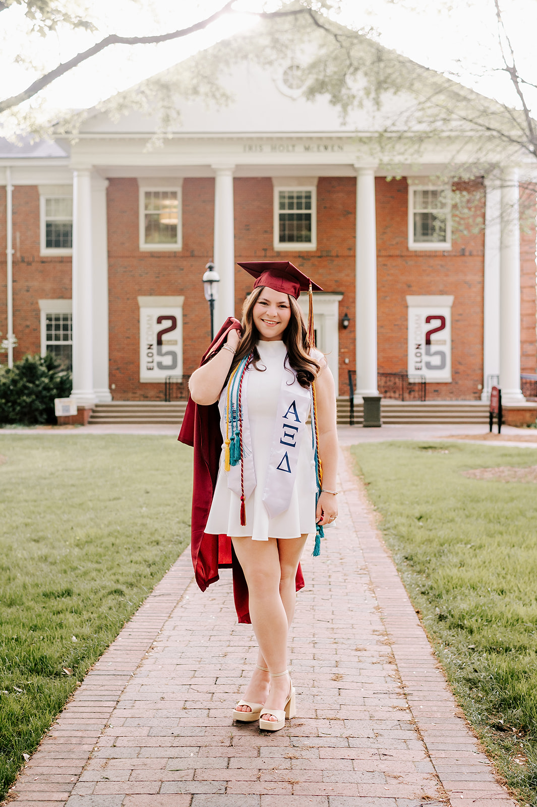 A happy grad walks a brick sidewalk in white dress and chords while carrying her gown