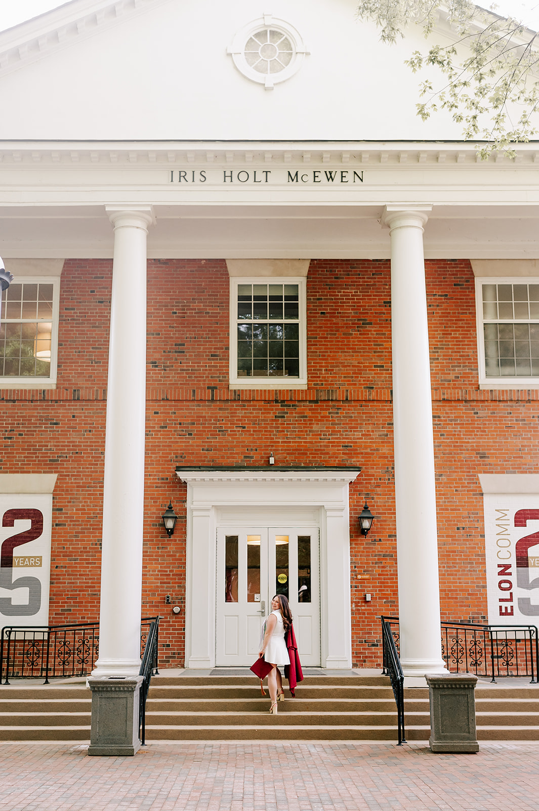 A grad walks un the steps of a classroom building in a white dress carrying her cap and gown