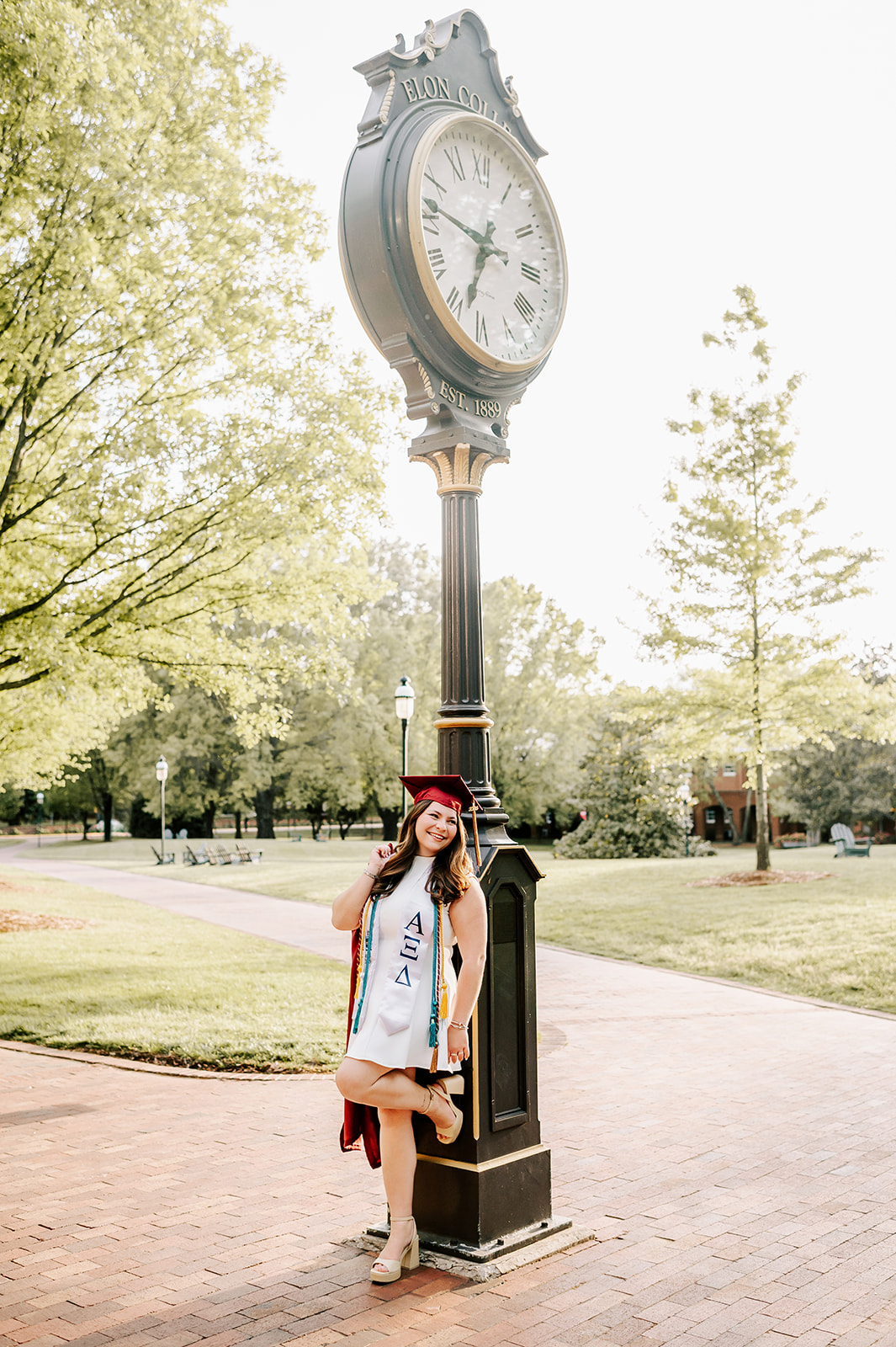 One of the excited Elon University graduates leans against the quad clock in a white dress and red cap