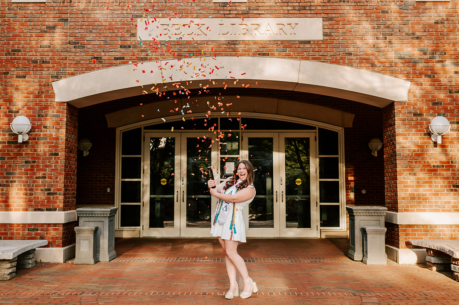 A happy grad pops a confetti cannon in a white dress with a big smile in front of Belk Library