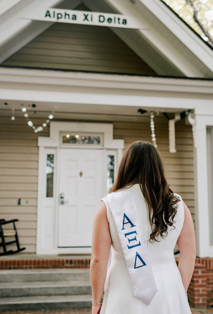 A Elon University graduate walks towards her sorority house porch in a white dress and stole