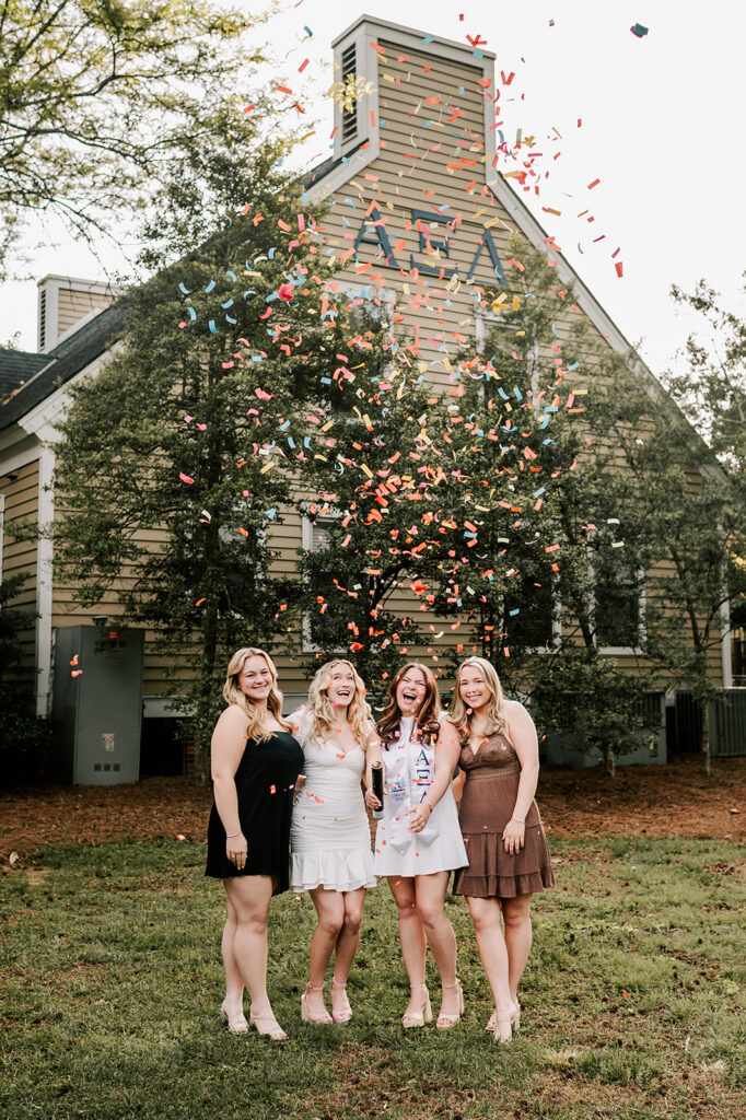 A group of sorority sisters laugh and pop confetti cannon in the lawn behind their house