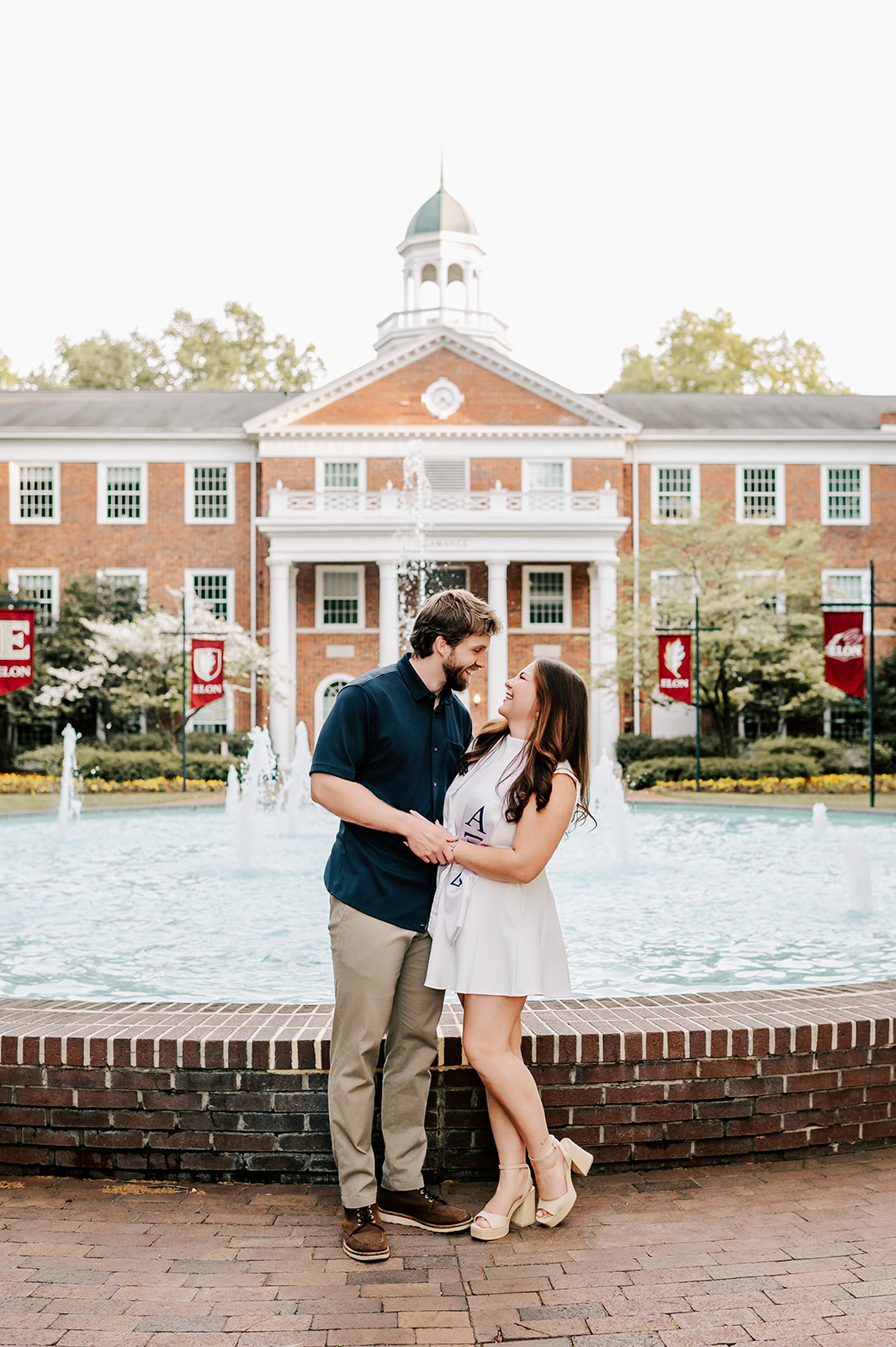 Laughing Elon University graduates embrace by a fountain and lean in for a kiss while standing and laughing