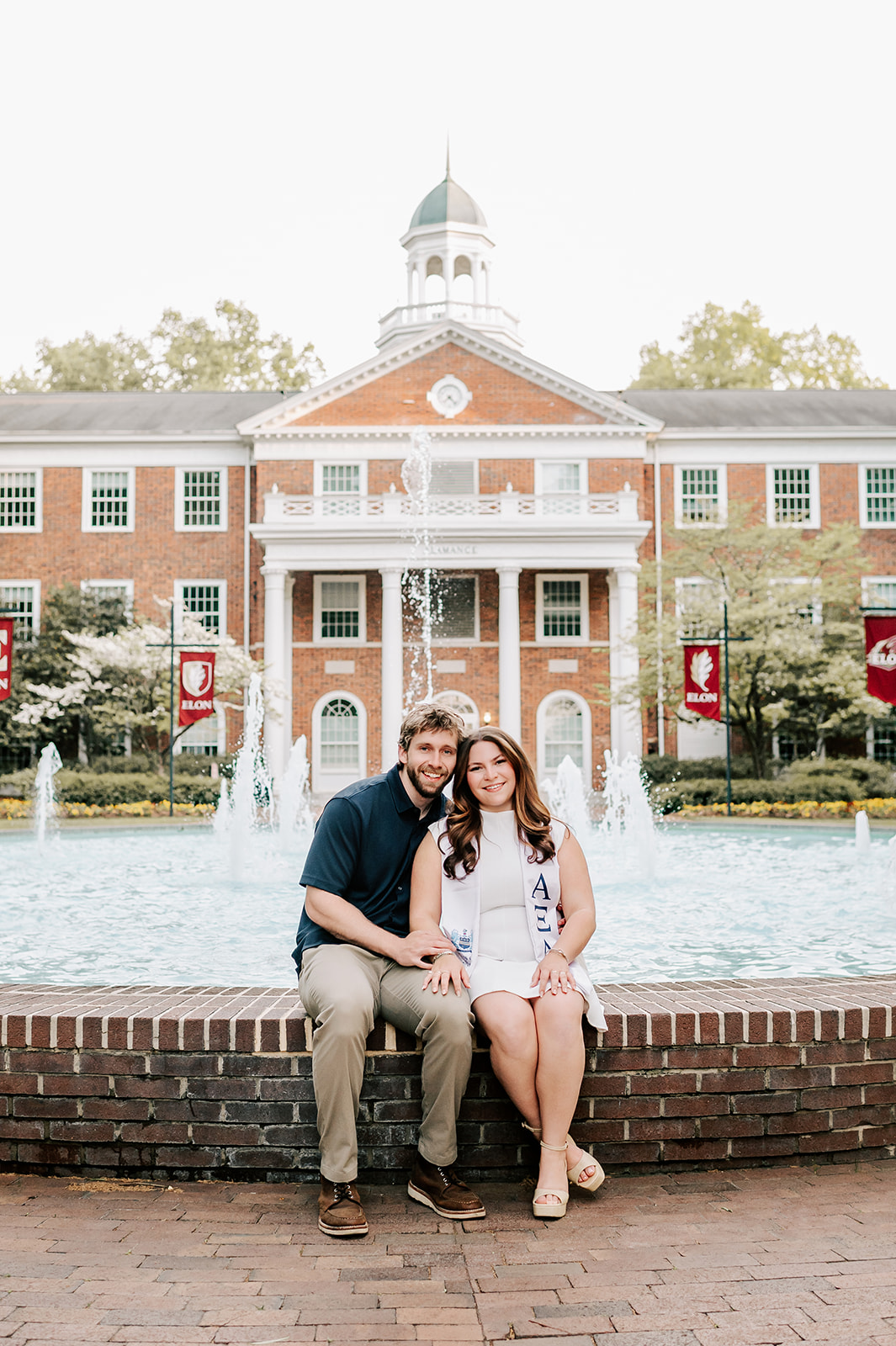Happy Elon University graduates sit on the edge of a fountain in white and blue and sorority rigalia