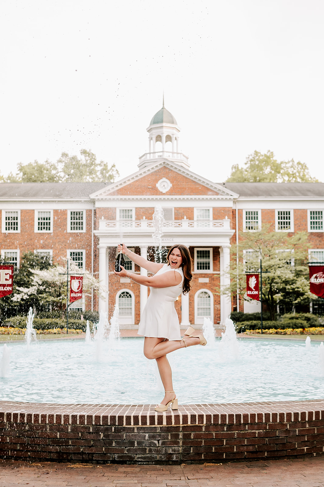 A woman celebrates with a bottle of champagne in a white dress on a fountain edge