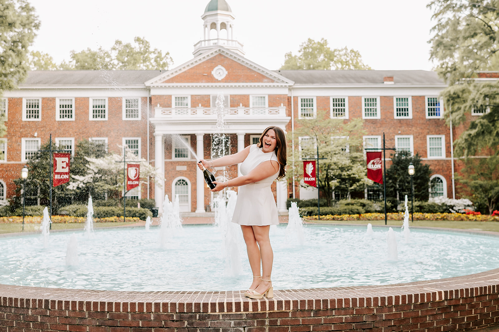 A happy woman pops and sprays a bottle of Champagne while standing on a fountain