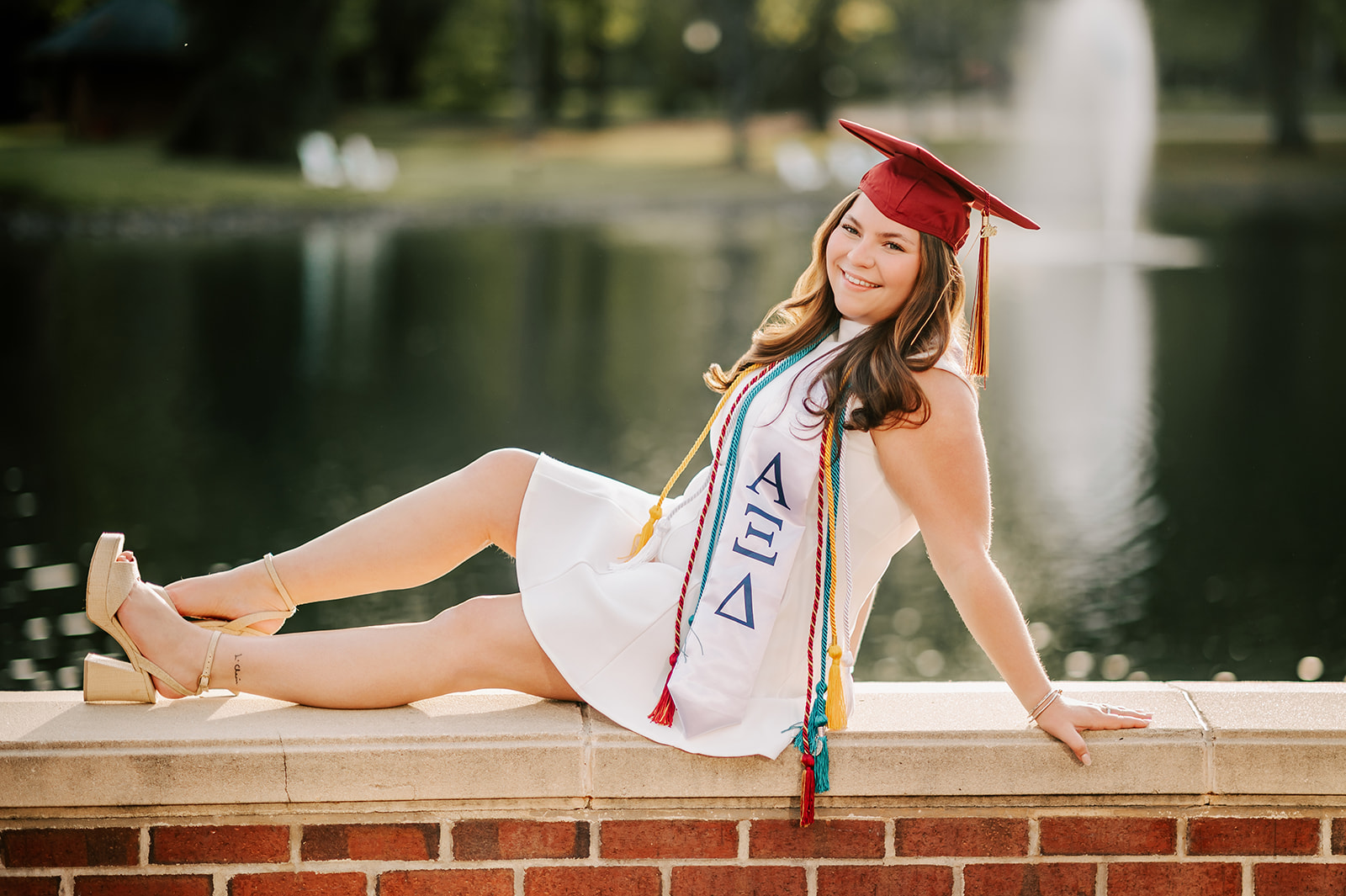One of the Elon University graduates in her cap and chords sits across a short wall by the lake smiling and leaning back