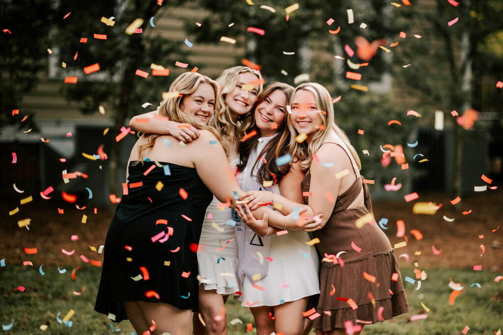A group of Elon University graduates hug under a shower of confetti in a lawn