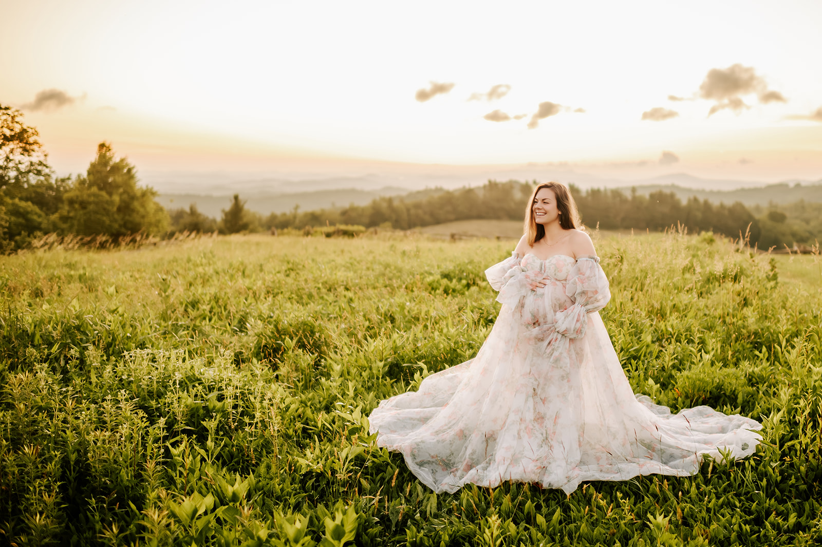 A laughing pregnant woman stands in a field of tall grass at sunrise on a mountain overlook