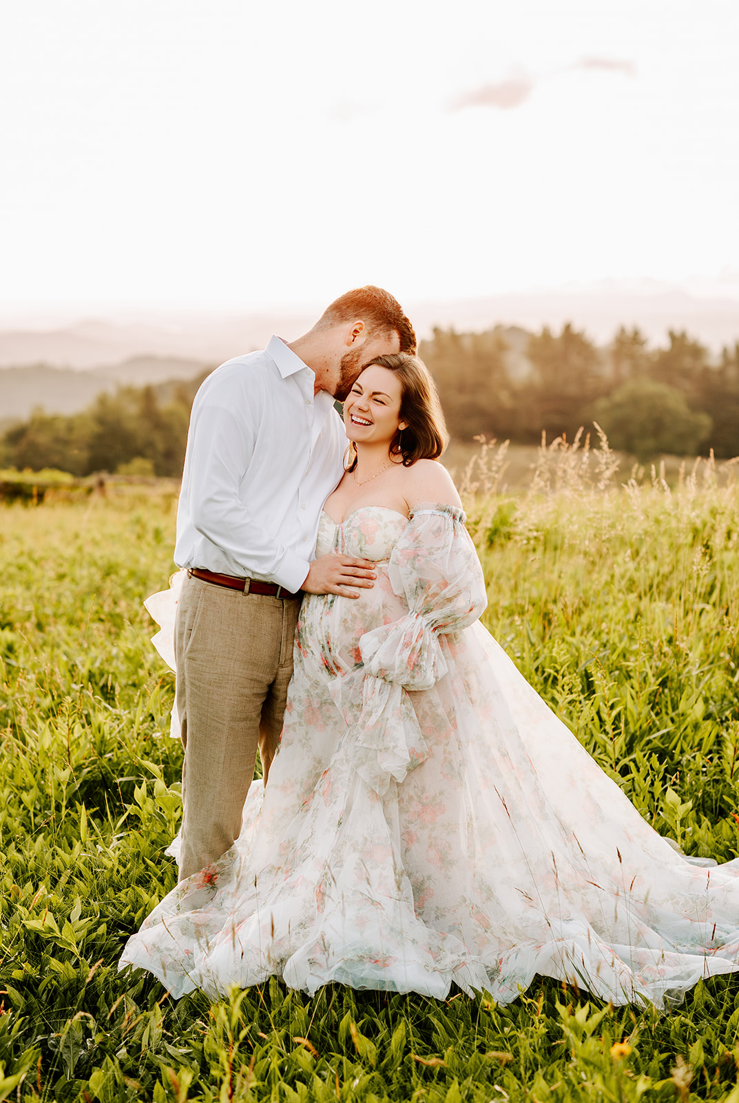 A mother to be laughs as her husband hugs her and whispers in her ear in a meadow at sunset in the mountains after meeting doulas in Greensboro, NC